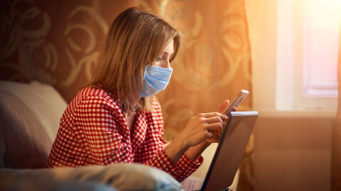 Junge Frau mit Maske und Smartphone sitzt vor einem Laptop Junge Frau mit Maske und Smartphone sitzt vor einem Laptop