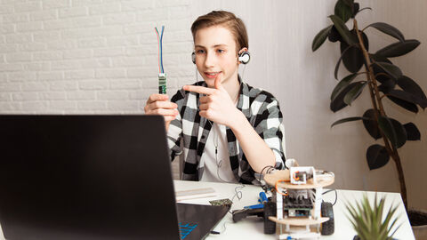 Junge sitzt am Computer und hält sein Physik Experiment in die Kamera