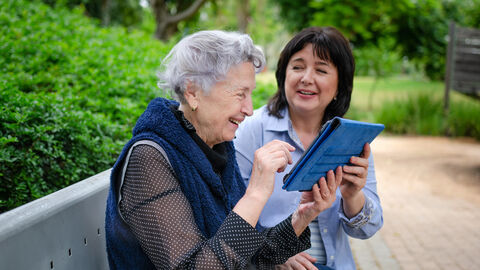 Eine Dame mittleren Alters und eine ältere Dame sitzen beisammen mit einem Tablet in Händen.