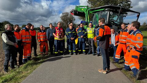 Viele Menschen stehen zusammen auf Straße zwischen Feldern. Im Hintergrund steht ein Traktor.
