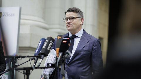 Ministerpräsident Rhein steht vor einer Ansammlung an Interview-Mikrofonen und spricht. Unter dem Arm hält er einen großen Aktenordner.