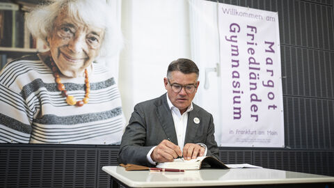 Ein Mann im Anzug sitzt an einem Tisch und schreibt in ein Buch, flankiert von einem großen Porträt von Margot Friedländer und einem Willkommensbanner des Margot-Friedländer-Gymnasiums.