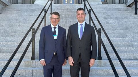Boris Rhein und Mike Needham stehen nebeneinander auf einer Treppe - im Hintergrund ist am Gebäude eine große Amerika Flagge befestigt