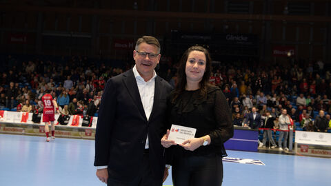 Ministerpräsident Boris Rhein steht auf dem Spielfeld der MT Melsungen. Die Ehrenamtliche Anja Scholtz hat von ihm eine Auszeichnung überreicht bekommen. Diese hält sie in der Hand.