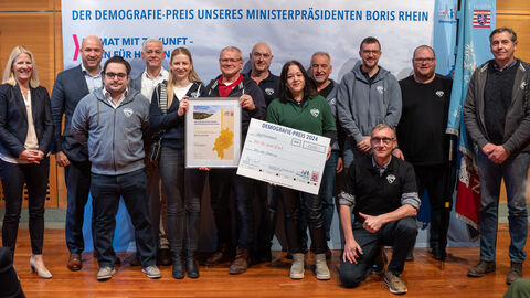 Eine Gruppe Personen steht vor einem Aufsteller mit der Aufschrift "Der Demografie-Preis unseres Ministerpräsidenten Boris Rhein". Zwei Personen halten eine gerahmte Urkunde in Händen, eine weitere einen Scheck.